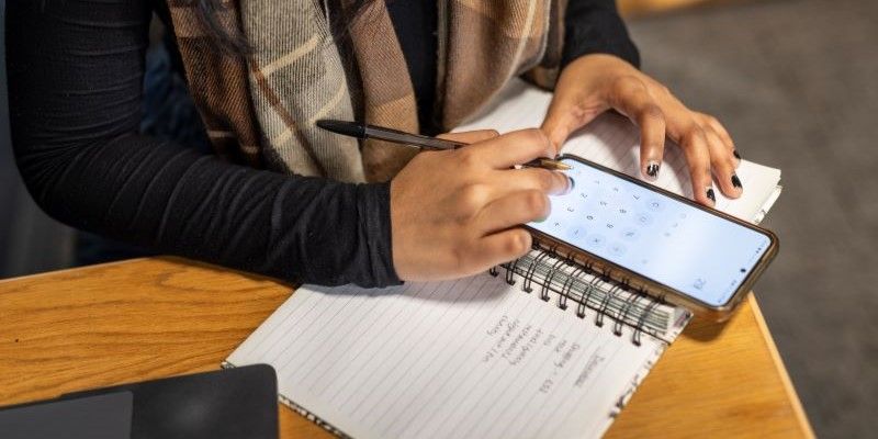 A student using a calculator app on their phone while holding a pen and taking notes on paper.