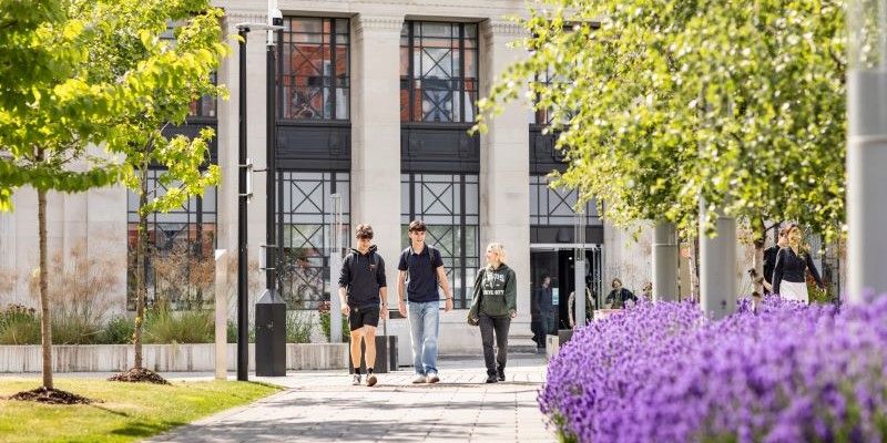 Three students walk toward the camera along a tree-lined path, smiling and engaged in conversation.