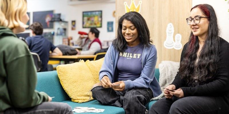 Three students sat on a sofa, smiling and playing a card game together.