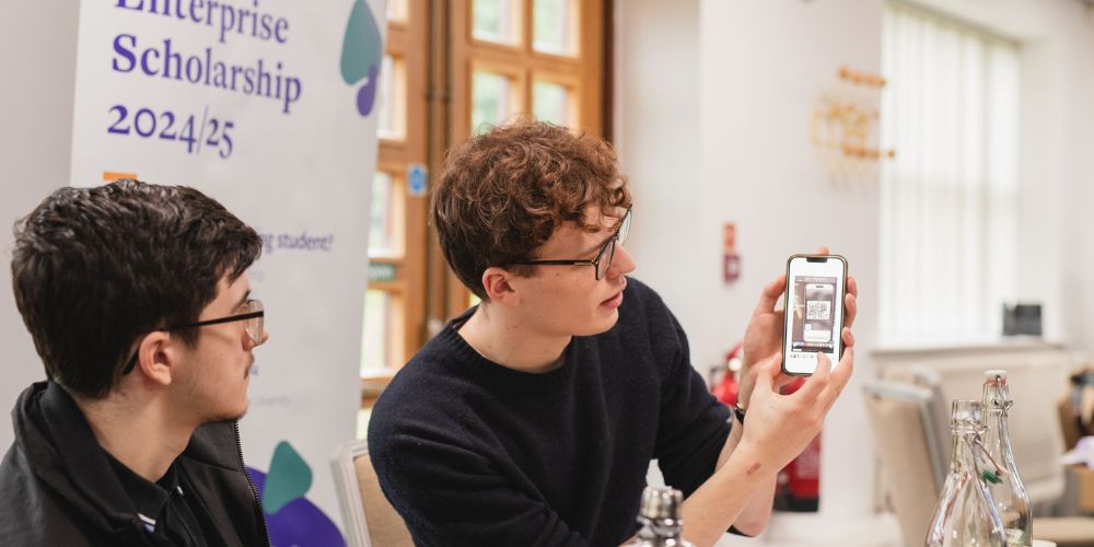 Two students sit at a table, in front of a banner that says 'Enterprise Scholarships 2024/25'. the student on the right is holding their phone up, showing the student on the left their screen.