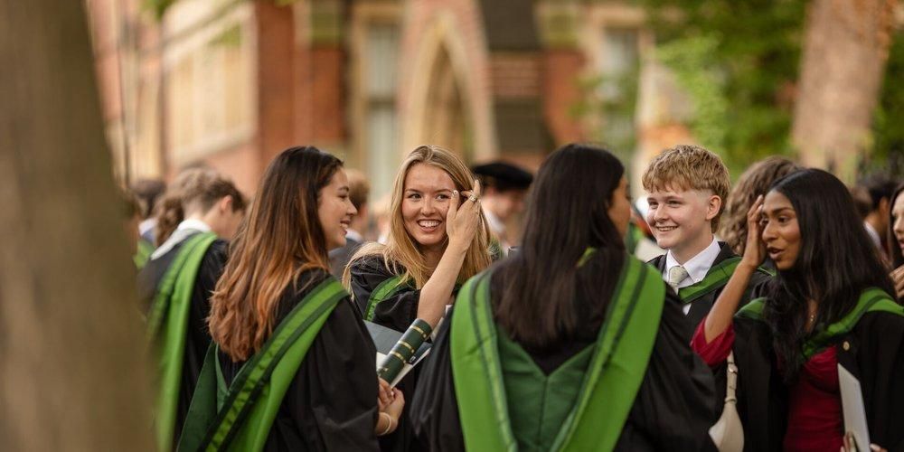 A group of graduates in black gowns and green hoods laugh and talk together outside a red brick building.