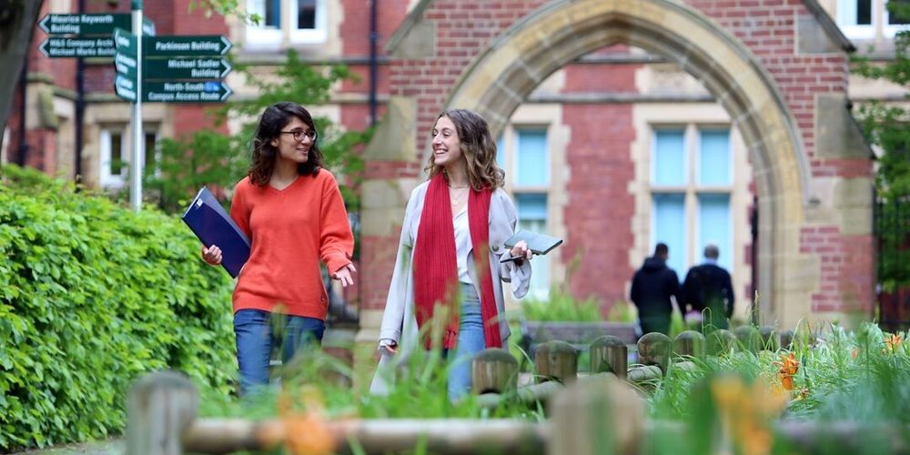 Two students are in conversation as they walk through a green space in front of a decorative red brick archway. They are carrying books and are smiling at each other.