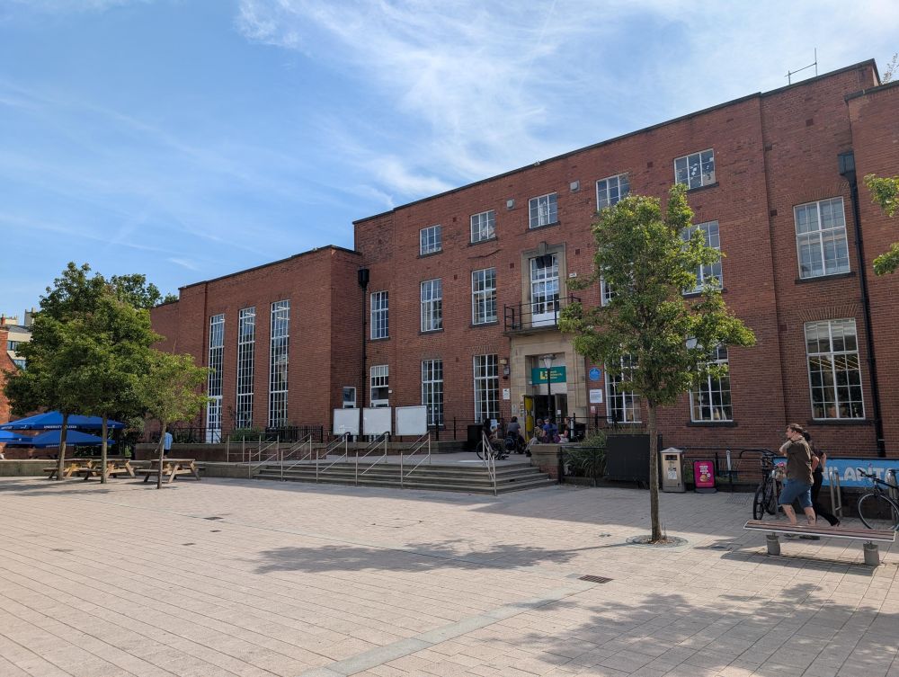 Red brick frontage of Leeds University Union across the open paving, with picnic benches to one side