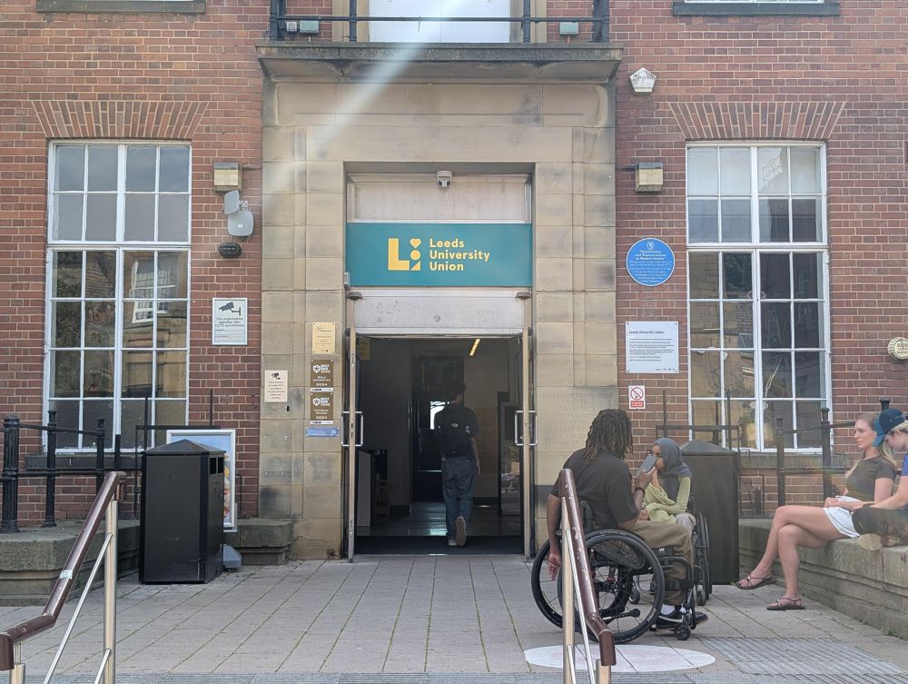 Main doors of Leeds University Union, with the teal and yellow sign above and a blue plaque alongside