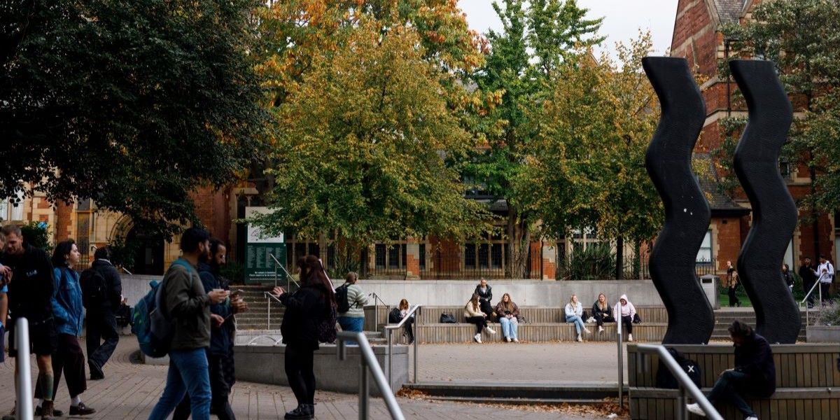 The trees outside the Great Hall building. Lush green and orange trees fill most of this image, with students walking and talking. The Sign For Art sculpture can be seen in the background.