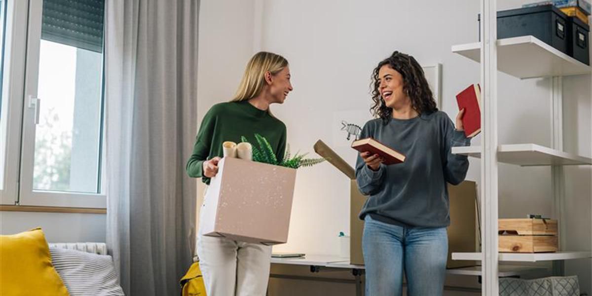 Two people stand in a bedroom, one is holding a box filled with various items like posters and a plant. The other is holding two books.