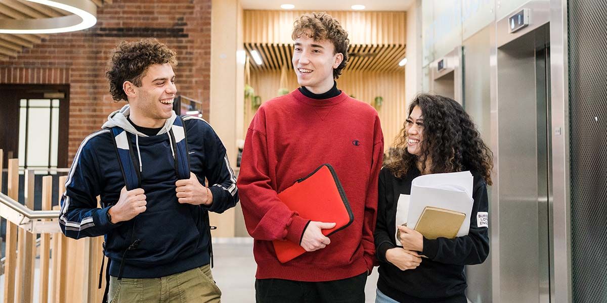 Three students walk along a corridor chatting and smiling in a modern building on campus.