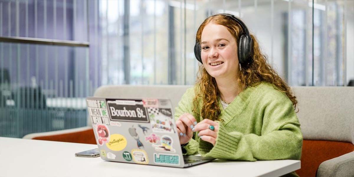 A student sat at a desk in a study space using a laptop and headphones and smiling.