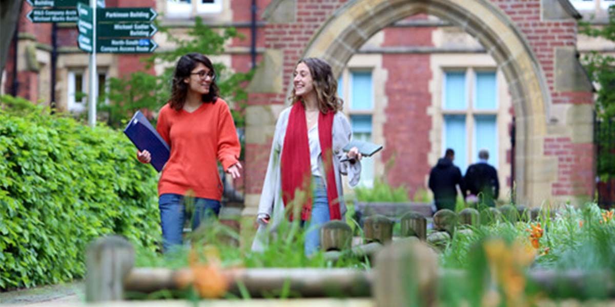 Two students are in conversation as they walk through a green space in Beech Grove Plaza. They are carrying books and are smiling at each other.