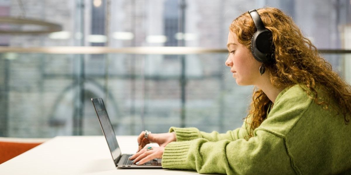 A side-on shot of a student sitting at a desk, typing on a laptop.  The student is wearing over-ear headphones and a green jumper.