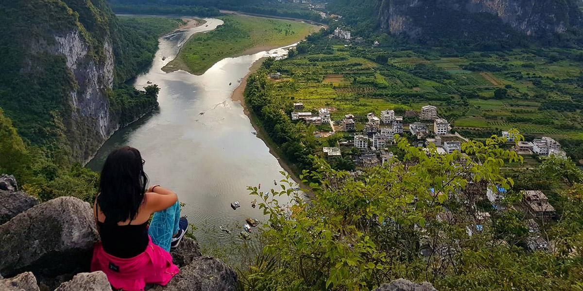 A study abroad student sits on a rock, facing over a valley with a river snaking through it.