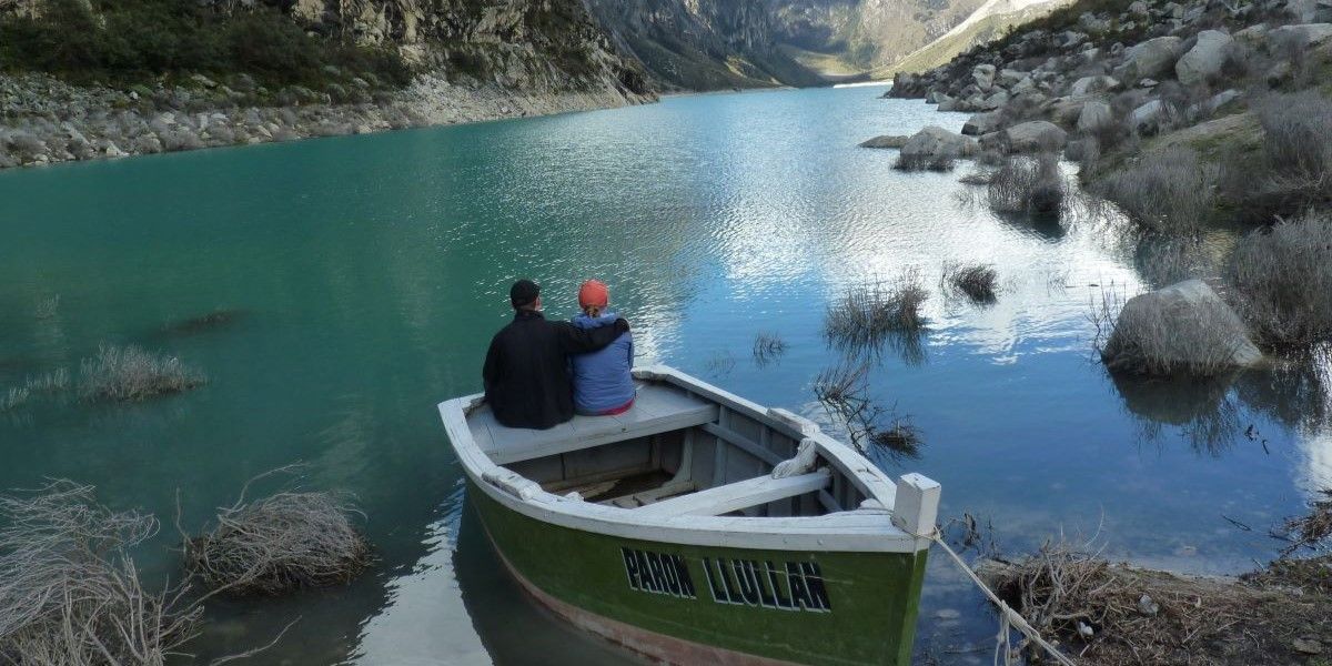 Two people sit on the edge of a wooden fishing boat, facing away from the camera, towards a big open blue lake, with rocks and hills either side. One has their arm around the other's shoulder as they sit peacefully.