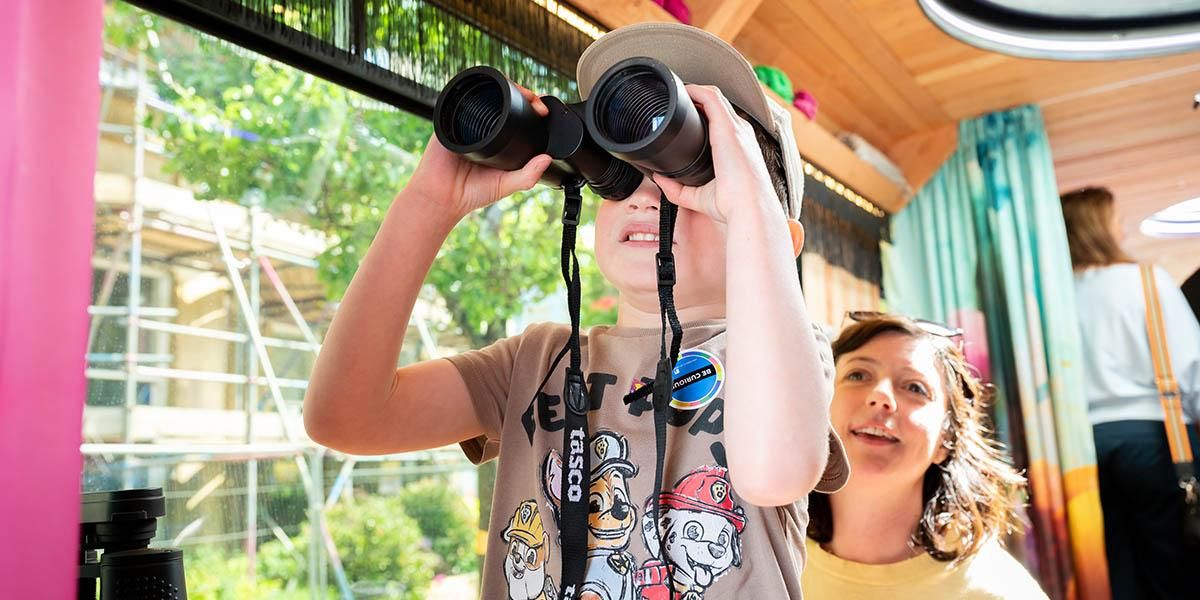 A child looks into the distance through large binoculars, while their parent sits behind, smiling.