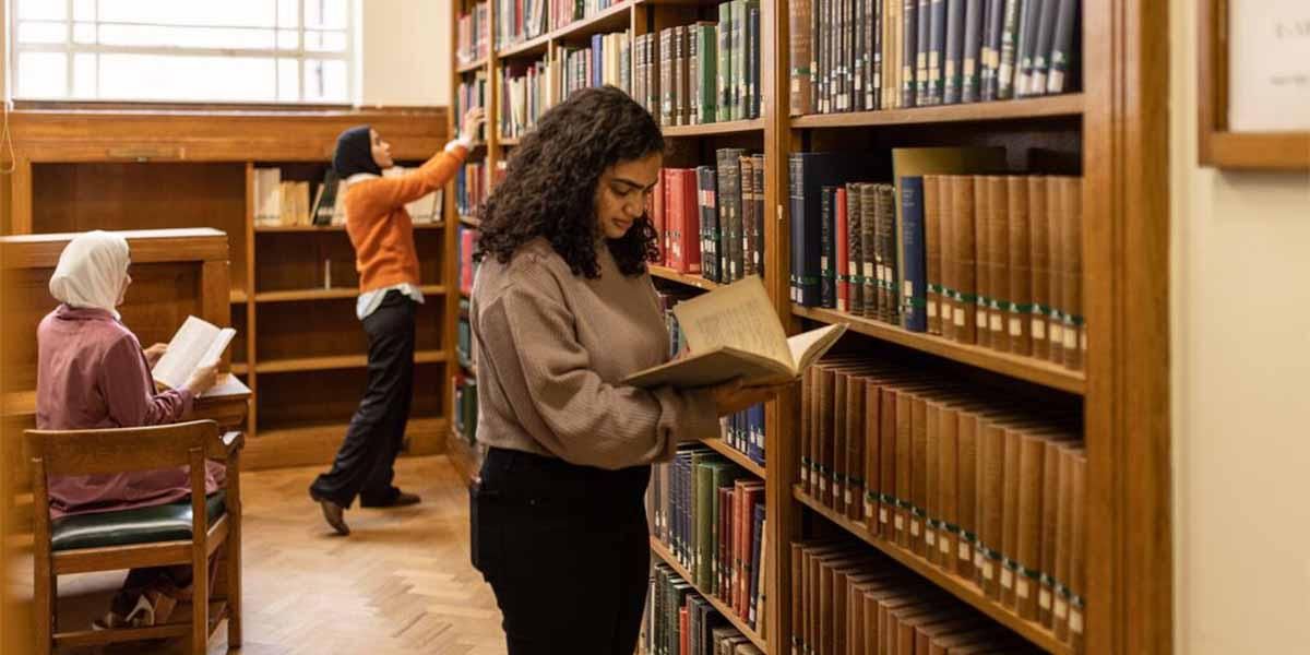 A student reading a book whilst stood next to the bookshelves on the ground floor of Brotherton Library.