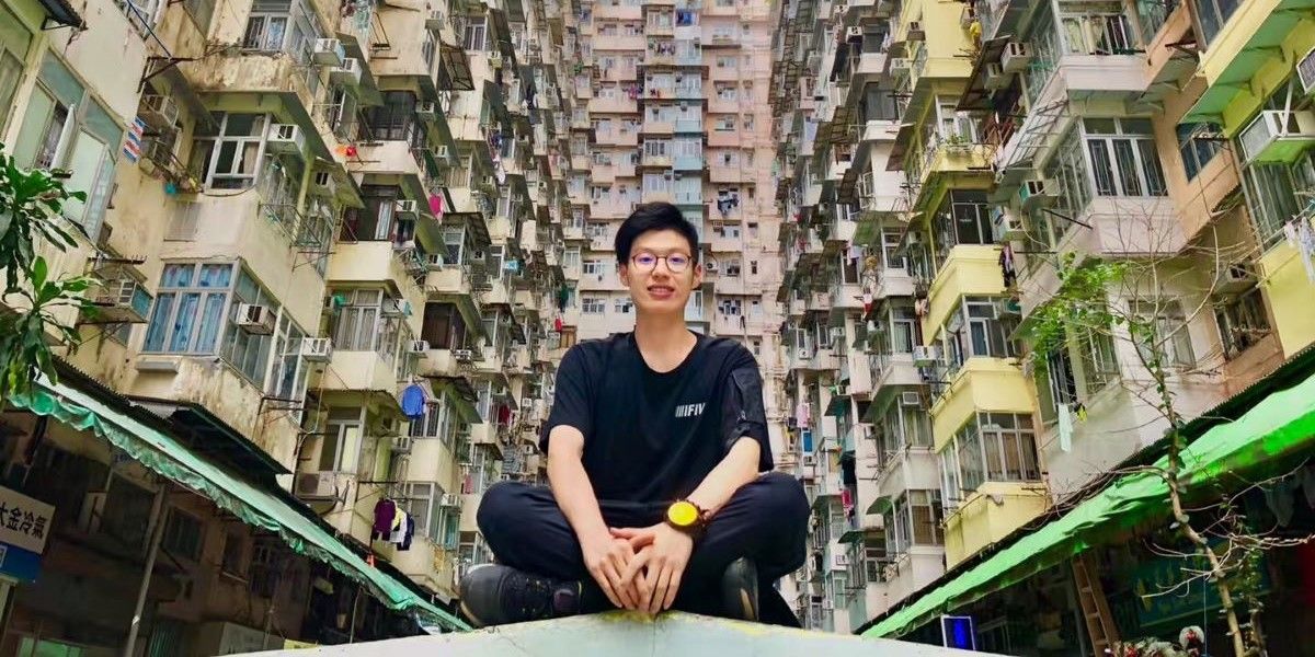 A study abroad student sits cross-legged on a roof, looking at the camera just below. Surrounding them are storeys of colourful Hong Kong apartments, wrapping around the streets on either side and behind.