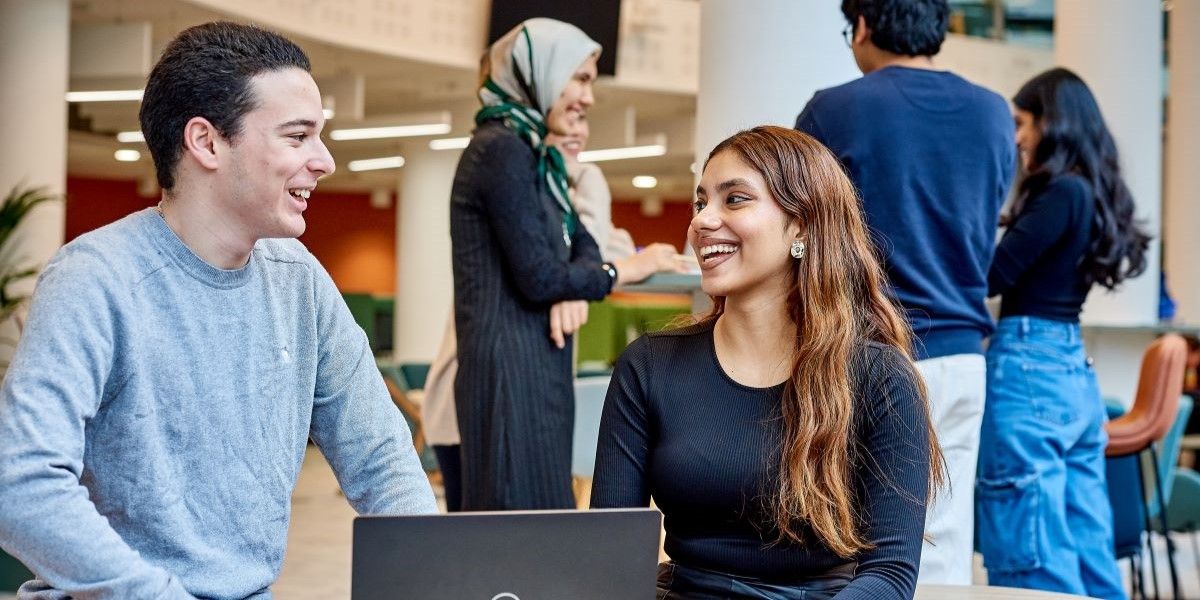 Two students sit at a small, round table and smile at each other and chat. There is a laptop in front of them on the table.