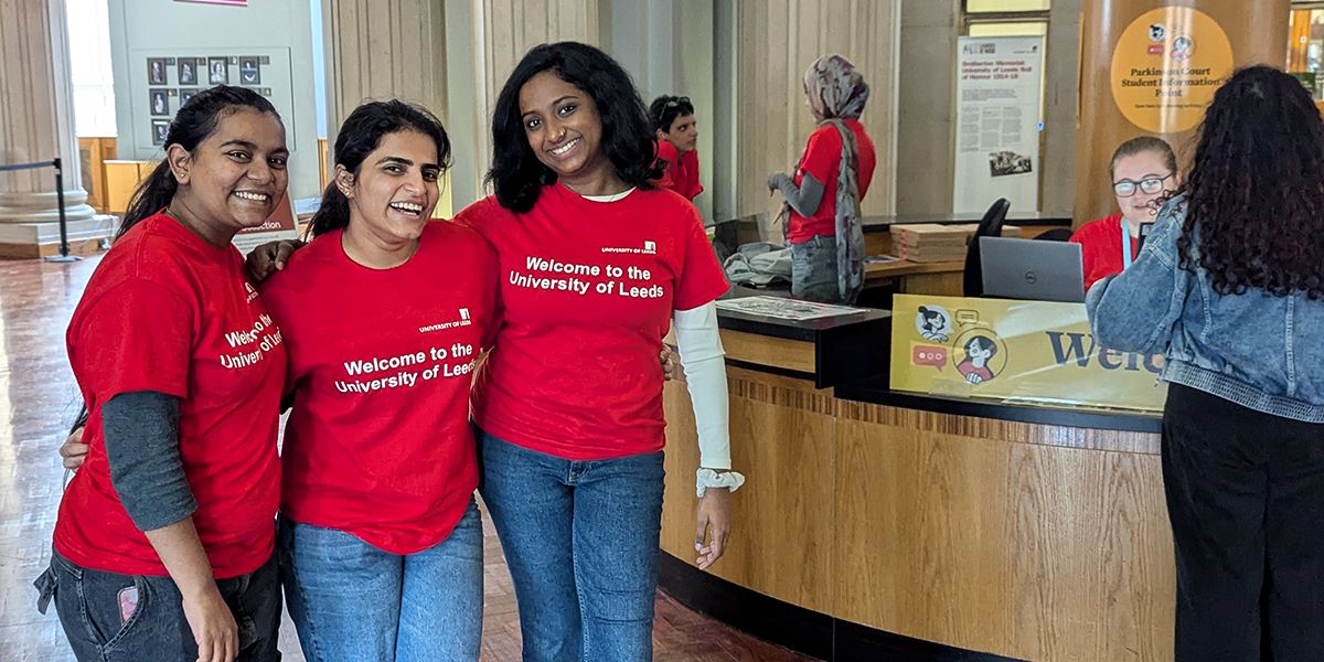 Three smiling student ambassadors wearing matching red "Welcome to the University of Leeds" t-shirts stand together inside a welcome hub building.