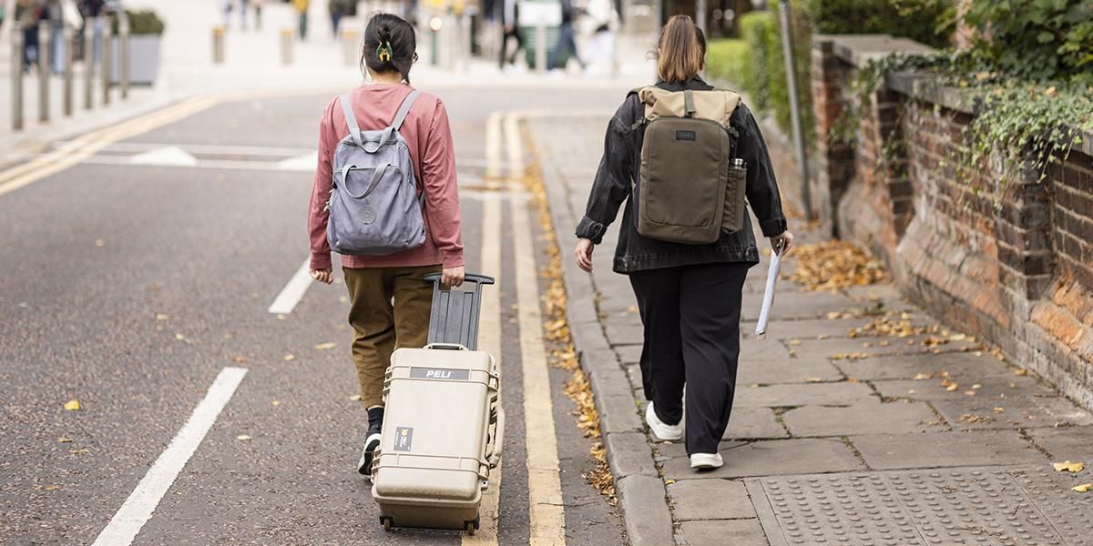 Two students walking on campus, one with a suitcase