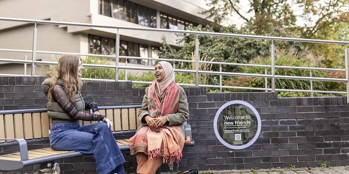 Two students talking on a bench next to a sign that says 'Welcome to new friends'