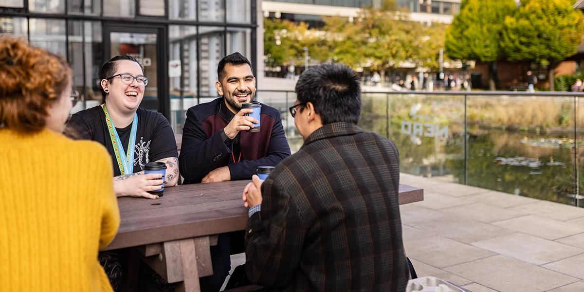 Students and a support worker drinking hot drinks on a bench by Roger Stevens pond