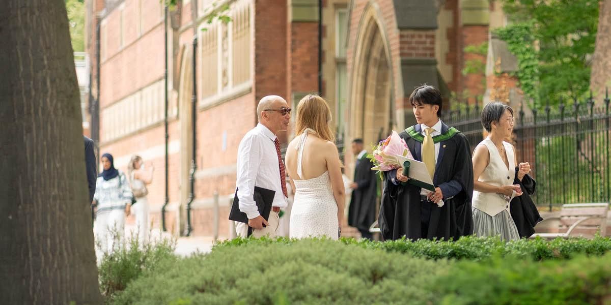 A graduate in a black gown and green hood holds flowers and a certificate folder while chatting with three guests outside a red-brick building on campus.