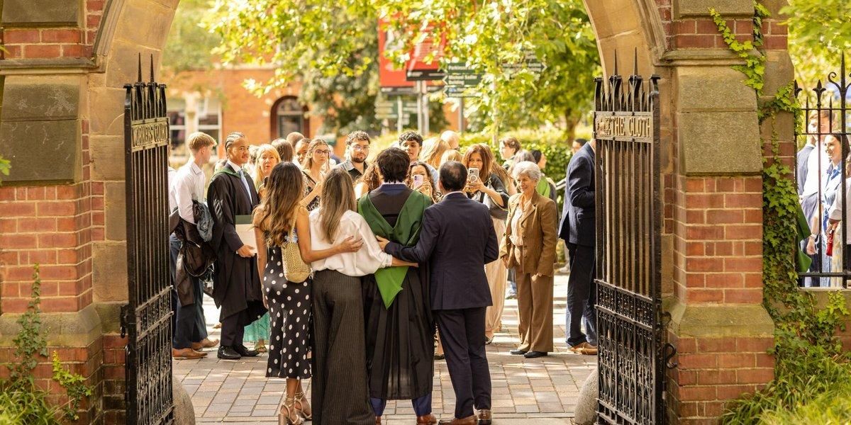 A graduate in a green hood stands arm in arm with family under a brick archway while friends take photos on their phones.