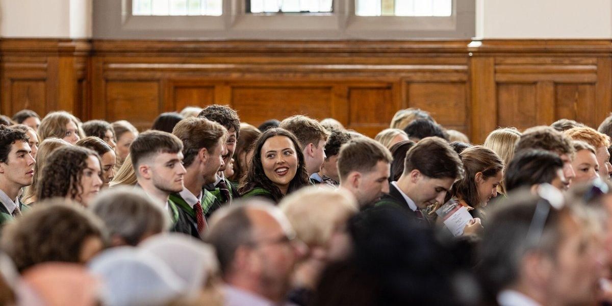 Rows of graduates in gowns chat before the ceremony, with wood panelling behind them and guests seated in the foreground.