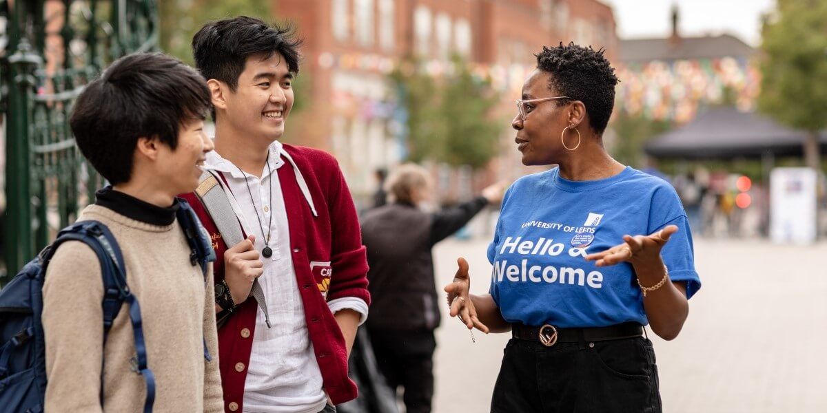 An ambassador in a blue t-shirt with the text 'Hello, Welcome' stands chatting to two smiling students, with the Leeds University Union building in the background.