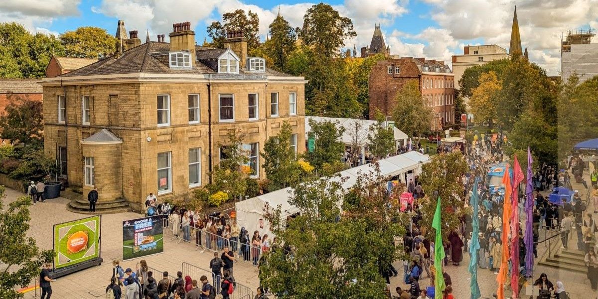 An overhead view of University Square during Welcome Week 2025. People queue for different stalls around the square and flags are waving. It's a sunny day with blue skies and fluffy white clouds.