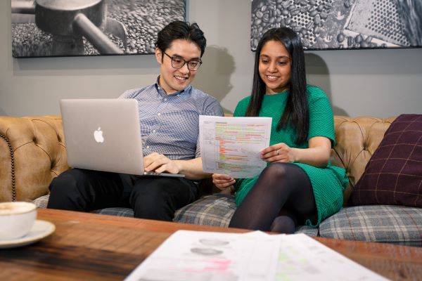 Two students sit on a sofa. One is using a laptop, the other is holding papers.