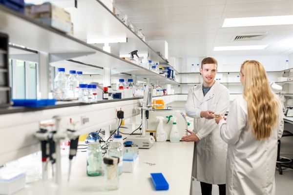 Two students work inside a laboratory. Both students wear white lab coats and one passes a small petri dish to the other, who is smiling.