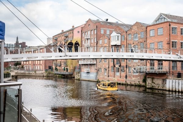 Four people stand on a bridge, looking over the river, with old factory buildings, now apartments, behind them.