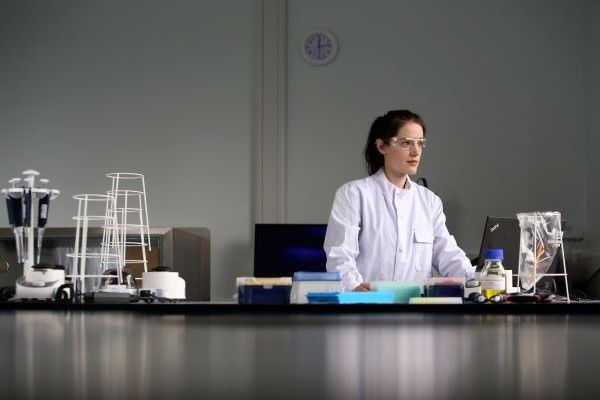 A student, wearing a white lab coat and protective glasses, works at a bench in a well-stocked laboratory. They're using a laptop to analyse the results from their experiment.