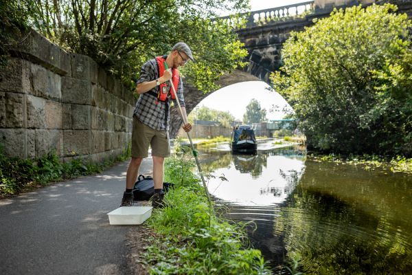 A postgraduate researcher stands studying the Leeds Liverpool canal, by the side of the canal bank while a canal barge goes by.