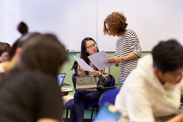 Landscape distant view of a tutor supporting a student. The tutor is stood and the student is sitting. The tutor is showing the student some papers and is dressed in a black and white striped top. There are several other students in the foreground and bac