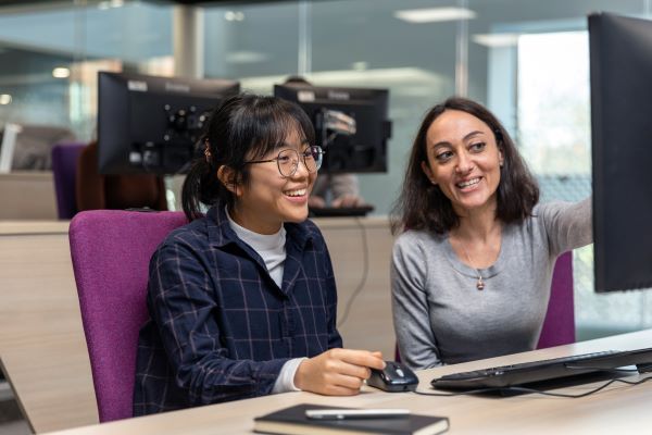 A students sits smiling at a computer while someone sits to their right, pointing to something on the computer screen.