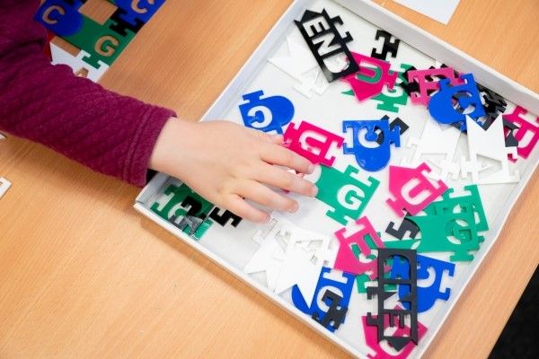 A child hovers their hand over a box containing a multi-coloured array of different letter shapes.