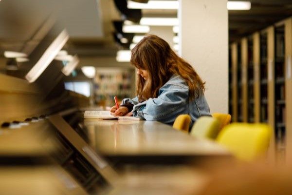A student is writing in a notebook with a textbook laid in front of them. They are studying inside a modern library, sat at a long, wooden desk.