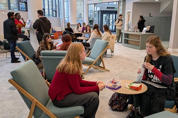 A group of people sit in a reception space, chatting, eating food and working on laptops.
