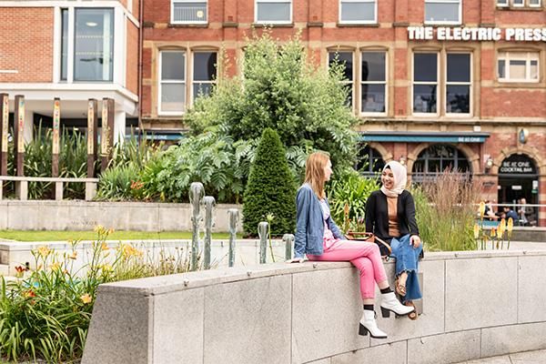 Long distant view of two students sitting and chatting on a stone wall in Nelson Mandela Gardens in central Leeds. One is wearing an orange top under a black cardigan and blue jeans with white headscarf, the second is wearing a lilac top under a blue shir