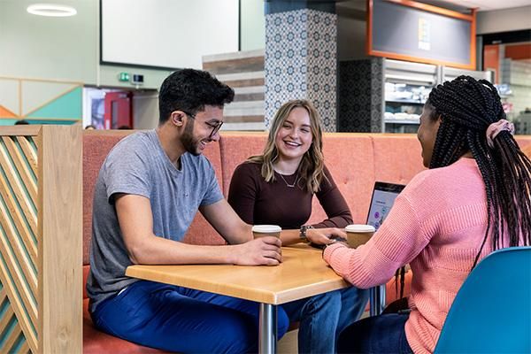 Three students sit at a wooden dining table in Common ground cafe. All three are in conversation and laughing. There is an open laptop on the table and all three are nursing paper coffee cups. One is wearing a blue t-shirt and jeans the second is wearing