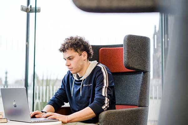 A student sits on a high-backed chair and works on their laptop inside one of the libraries on campus during the day.