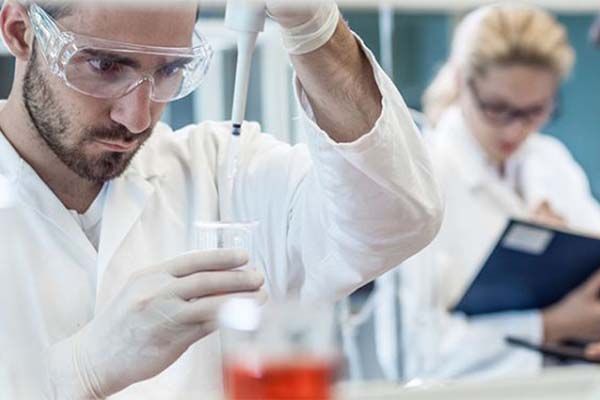 A student works in a lab carefully adding liquid to a beaker. Another person is working in the lab behind them.