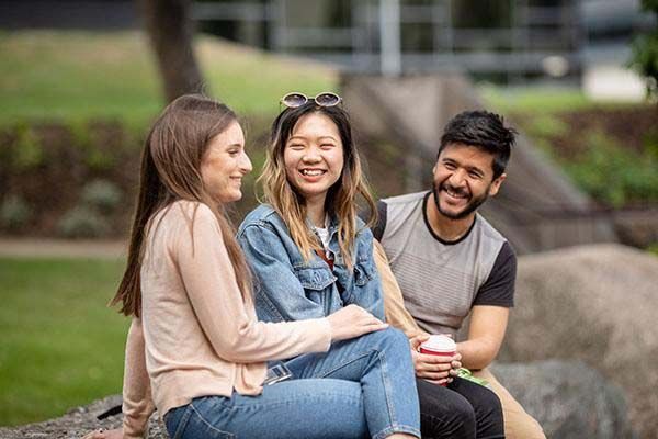 Three international students laughing whilst sitting on a rock on campus