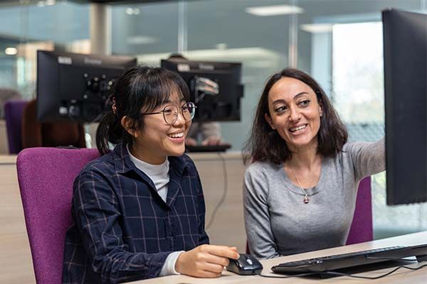 A tutor providing technical support to a student whilst sat at a desk working on a computer