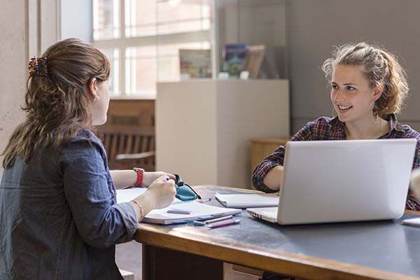 Students sat at a desk in the Parkinson Court with a laptop, chatting.