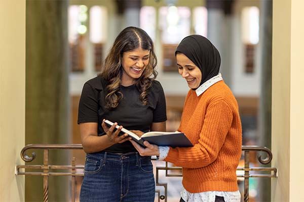 Two students in the library standing up and looking through a book together. Both students are smiling.