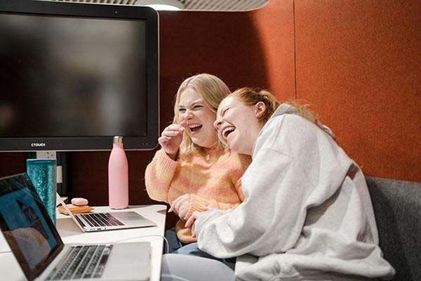 Two students lean against each other and laugh whilst working on their laptops inside a booth.