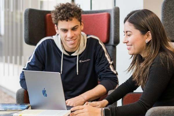 Two students sitting by a coffee table as they work together on a laptop. Both students are smiling.