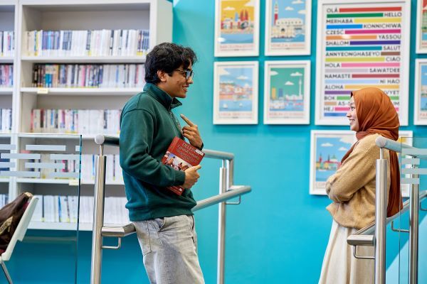 Two students stand and smile at each other as they chat. One of them is holding a thick, red textbook.